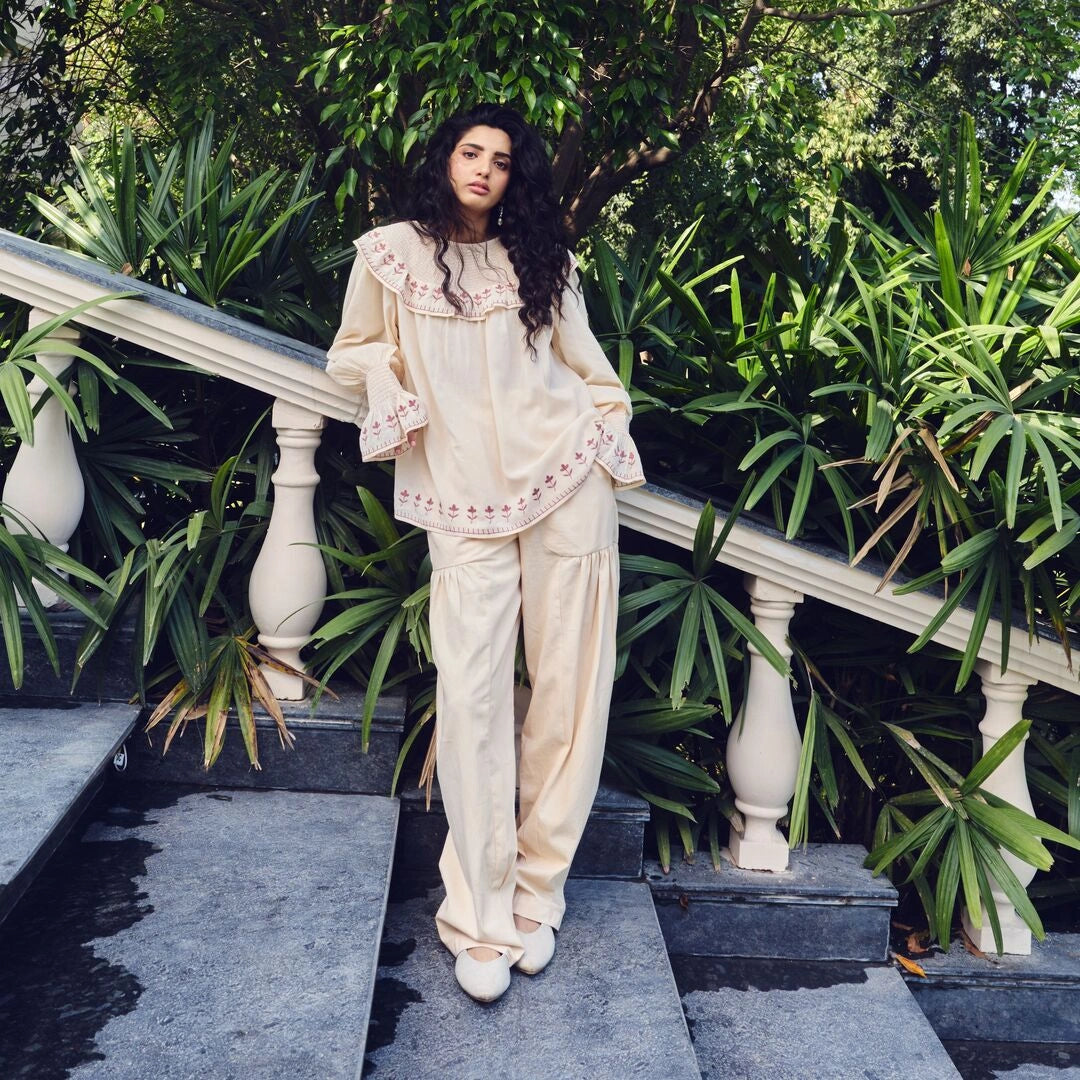 A woman stands on outdoor stone steps surrounded by lush green plants, wearing a cream-colored two-piece lounge set with delicate pink embroidery and ruffled details.