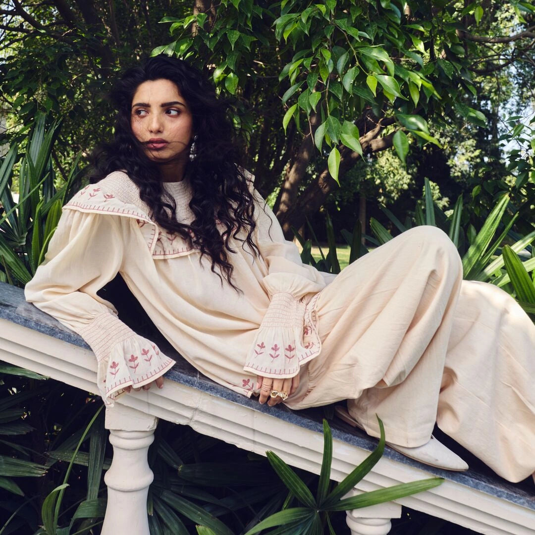 Woman in a cream-colored outfit sitting on a stone bench with greenery in the background