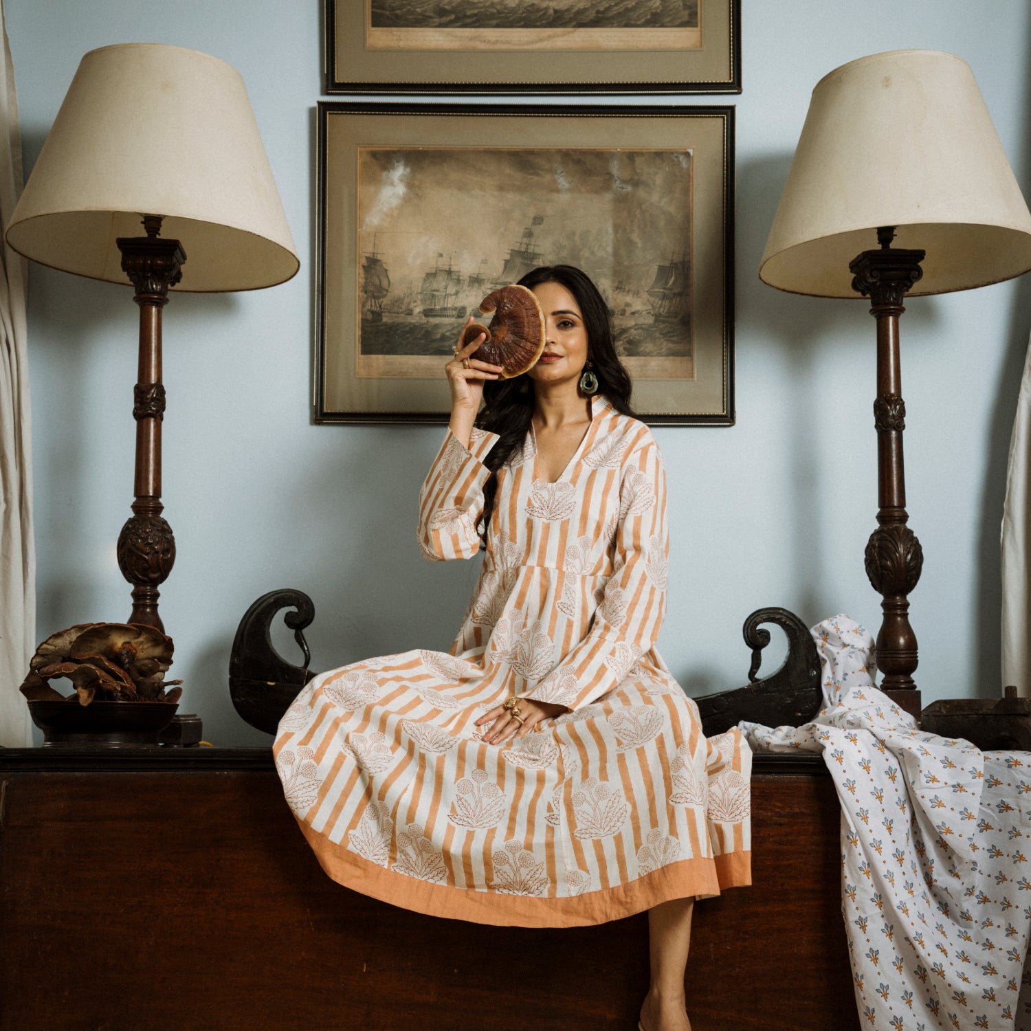Woman in a patterned dress sitting on a bench with lamps and framed pictures in the background.