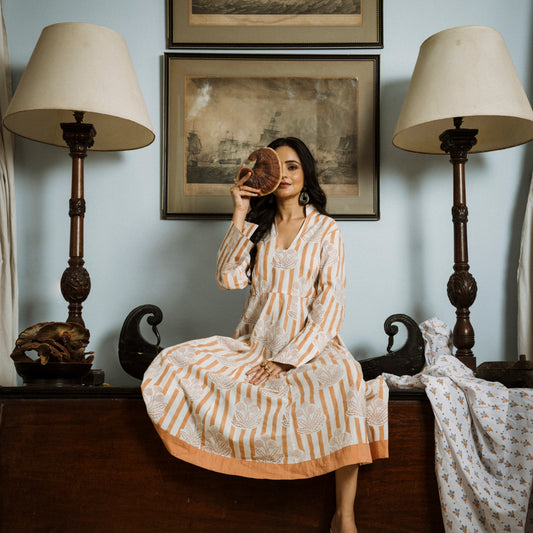 Woman in a patterned dress sitting on a bench with lamps and framed pictures in the background.