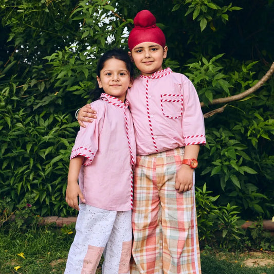 Two children in traditional pink and plaid outfits standing in a natural setting.