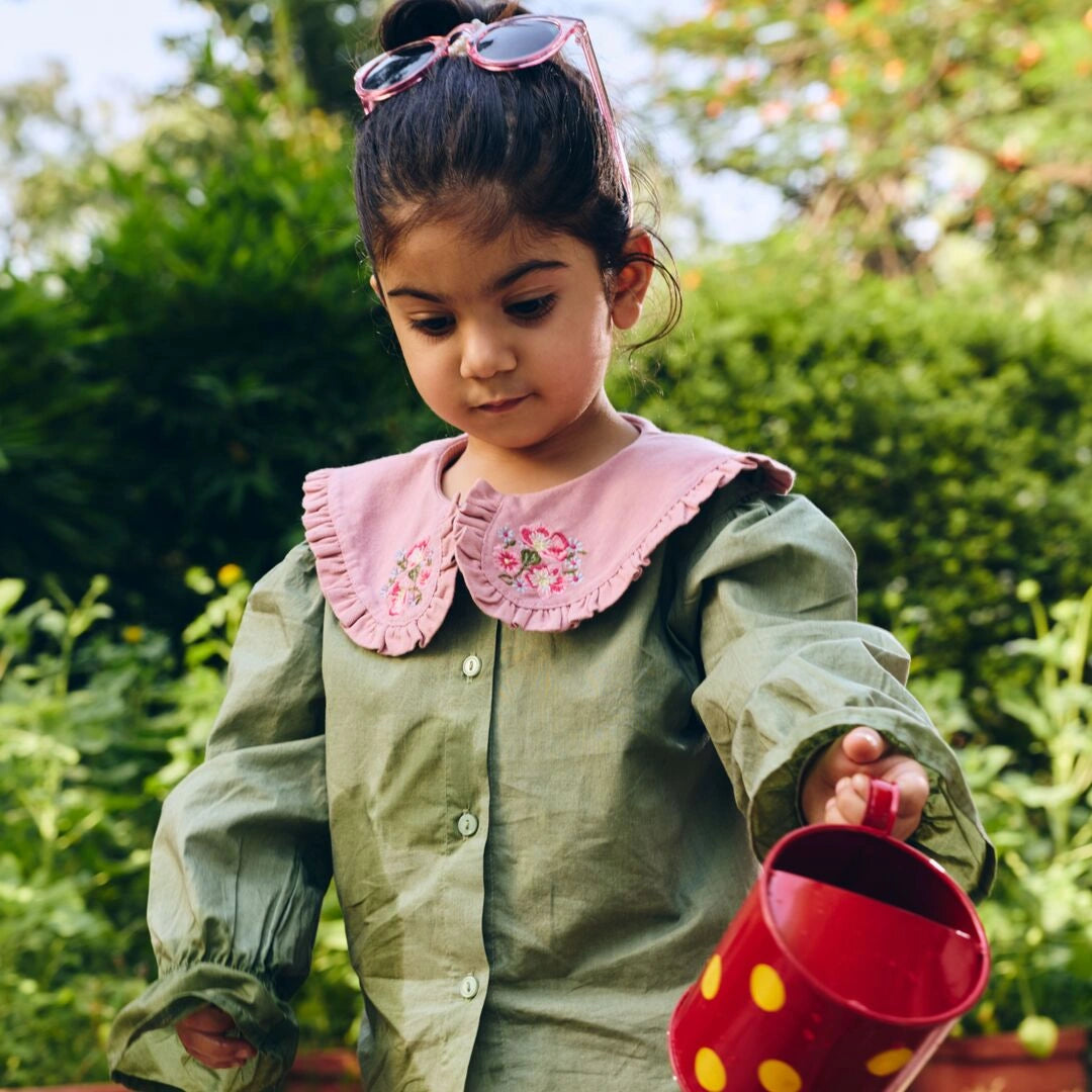 Child in a green raincoat with a pink collar holding a red watering can outdoors.