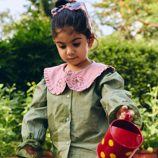 Child in a green raincoat with a pink collar holding a red watering can outdoors.