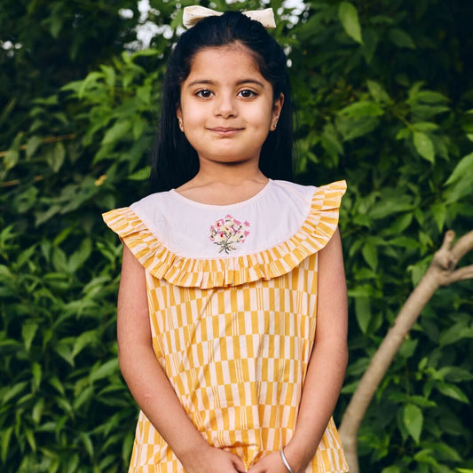 Young girl wearing a yellow checkered dress with a floral design in a green outdoor setting